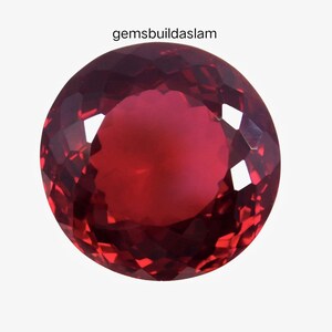 Può includere: Una pietra preziosa rotonda e sfaccettata di colore rosso intenso con una superficie brillante e riflettente. Il testo gemsbuildaslam è stampato in nero sopra la pietra. La pietra mostra un complesso motivo di tagli geometrici.