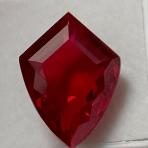 May include: A faceted, deep red gemstone with a unique shield-like shape. The gem is set against a white background, highlighting its brilliant cut and rich colour. This gemstone is ideal for jewellery making.