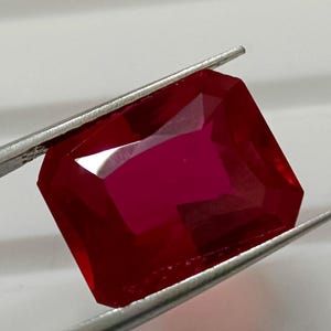 Può includere: Un rubino vibrante, taglio rettangolare, tenuto da pinzette d'argento. Il rubino mostra un colore rosso intenso e ricco con molteplici sfaccettature che riflettono la luce. La forma della gemma è un taglio smeraldo classico.