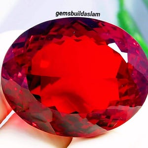 Könnte beinhalten: Ein großer, oval geschliffener, leuchtend roter Edelstein mit komplexen Facetten, die das Licht reflektieren. Der Stein hat einen tiefen, gesättigten karmesinroten Farbton und ist mit dem Text gemsbuildaslam über dem oberen Rand versehen.