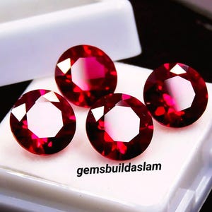 May include: Four round, faceted, deep red gemstones arranged on a white display base. The stones exhibit a brilliant cut with multiple reflective surfaces. The text "gemsbuildaslam" is printed in black on the white surface near the bottom centre.