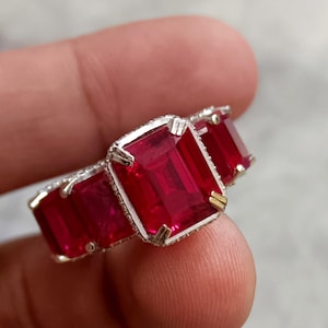 Puede incluir: Primer plano de un anillo de plata con tres grandes piedras preciosas rojas rectangulares. El engaste del anillo tiene un diseño detallado, y las piedras preciosas son el punto focal de la joya.