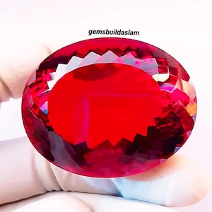 May include: A large, oval-cut, vibrant red gemstone with a faceted surface that reflects light. The stone features a brilliant, deep crimson hue throughout its structure. The text "gemsbuildaslam" is printed in black above the gemstone.