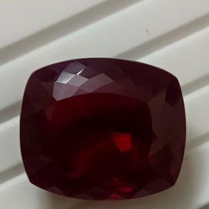 Könnte beinhalten: Ein facettierter Rubin-Edelstein im Kissenschliff. Der Edelstein hat eine tiefe, satte rote Farbe mit mehreren Facetten, die das Licht reflektieren. Die Form ist quadratisch mit abgerundeten Ecken.