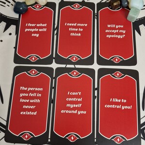 Puede incluir: Seis cartas de tarot con caras rojas y bordes negros, cada una con texto blanco. Las frases incluyen "Temo lo que dir&aacute; la gente" y "&iexcl;Me gusta controlarte!". Varias piedras preciosas se colocan cerca de las cartas.