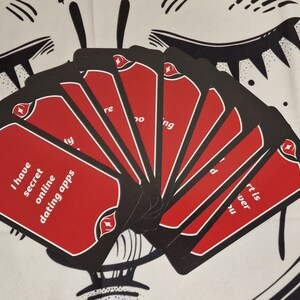 Puede incluir: Un mazo de cartas negras y rojas en abanico, con texto en cada carta. Las cartas tienen un fondo rojo con texto blanco y un borde negro. El texto en una carta dice: "Tengo aplicaciones de citas en l&iacute;nea secretas".