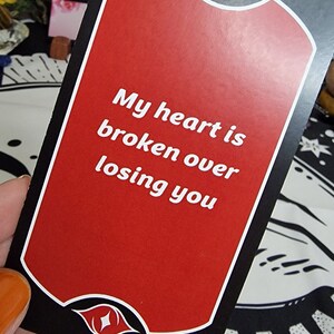 Puede incluir: Una carta de tarot con fondo rojo y borde negro. La carta presenta el texto "My heart is broken over losing you" en blanco. Dise&ntilde;os decorativos de ojos y labios en la parte superior e inferior.