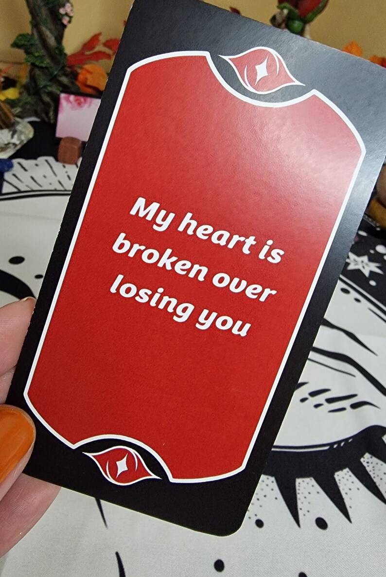 Puede incluir: Una carta de tarot con fondo rojo y borde negro. La carta presenta el texto "My heart is broken over losing you" en blanco. Dise&ntilde;os decorativos de ojos y labios en la parte superior e inferior.