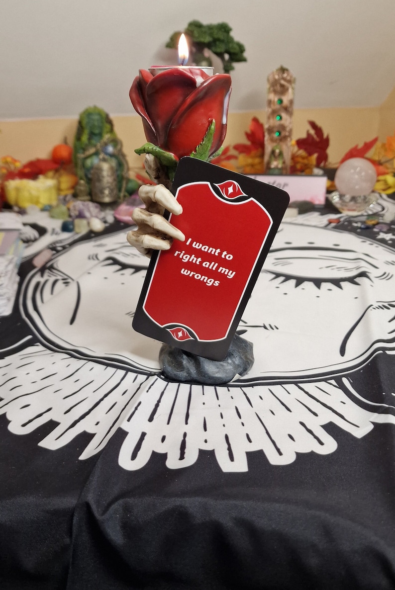 Puede incluir: Una carta de tarot roja y negra con el texto "I want to right all my wrongs" es sostenida por una mano de esqueleto. La carta est&aacute; colocada sobre una tela estampada en blanco y negro. Un candelabro de rosa roja con una vela encendida est&aacute; en el fondo.