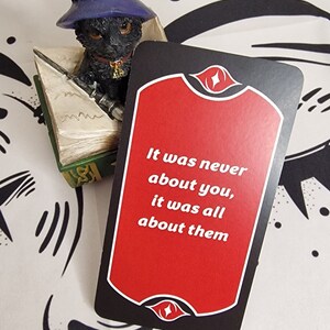 Puede incluir: Figura de gato negro con sombrero de mago azul, sentado sobre un libro en miniatura, junto a una tarjeta con el texto "It was never about you, it was all about them" sobre fondo rojo.