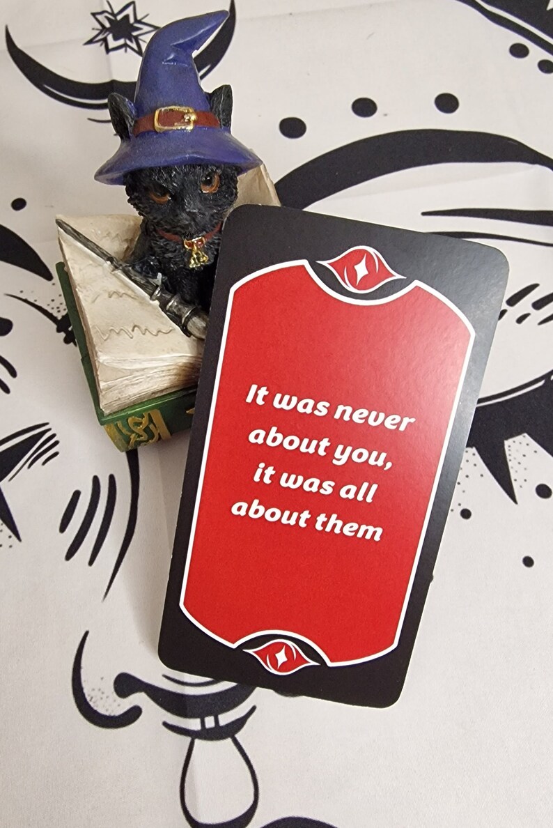 Puede incluir: Figura de gato negro con sombrero de mago azul, sentado sobre un libro en miniatura, junto a una tarjeta con el texto "It was never about you, it was all about them" sobre fondo rojo.