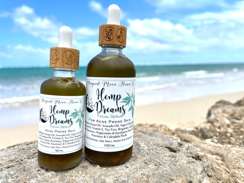 May include: Two glass bottles of Hemp Dreams facial serum for acne prone skin. The bottles are green and have a wooden top with a dropper. The bottles are sitting on a rock with a beach and ocean in the background.
