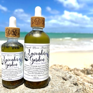 May include: Two glass bottles of Lavender Garden facial serum, one 50 ml and one 100 ml, with wooden caps and labels. The bottles are sitting on a rock with a beach and ocean in the background.