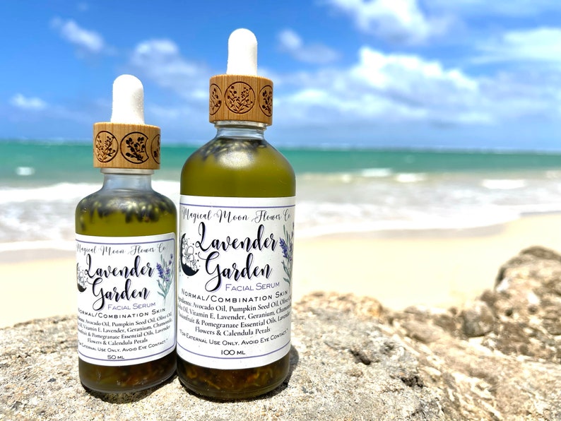 May include: Two glass bottles of Lavender Garden facial serum, one 50 ml and one 100 ml, with wooden caps and labels. The bottles are sitting on a rock with a beach and ocean in the background.