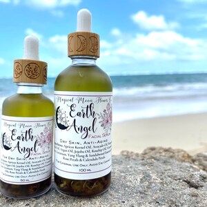 May include: Two glass bottles of facial serum with dropper tops and wooden caps. The bottles are labeled "Magical Moon Flowers Co. Earth Angel Facial Serum". The larger bottle is labeled "Dry Skin, Anti-Aging" and contains 100 ml. The smaller bottle is labeled "For Dry Skin, Anti-Aging" and contains 50 ml. The bottles are sitting on a rock with a beach and ocean in the background.