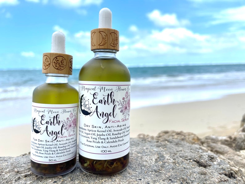May include: Two glass bottles of facial serum with dropper tops and wooden caps. The bottles are labeled "Magical Moon Flowers Co. Earth Angel Facial Serum". The larger bottle is labeled "Dry Skin, Anti-Aging" and contains 100 ml. The smaller bottle is labeled "For Dry Skin, Anti-Aging" and contains 50 ml. The bottles are sitting on a rock with a beach and ocean in the background.