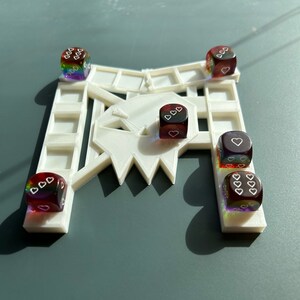 May include: A white plastic game board with a rainbow and red dice with white hearts on each side. The dice are on the board in different positions.