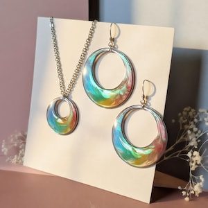 K&ouml;nnte beinhalten: Ein Schmuckset bestehend aus einer Halskette und zwei passenden Creolen. Jedes Schmuckst&uuml;ck verf&uuml;gt &uuml;ber einen kreisf&ouml;rmigen Metallrahmen mit einer wirbelnden, mehrfarbigen Harzeinlage in leuchtenden Blau-, Gr&uuml;n-, Gelb- und Rosat&ouml;nen.