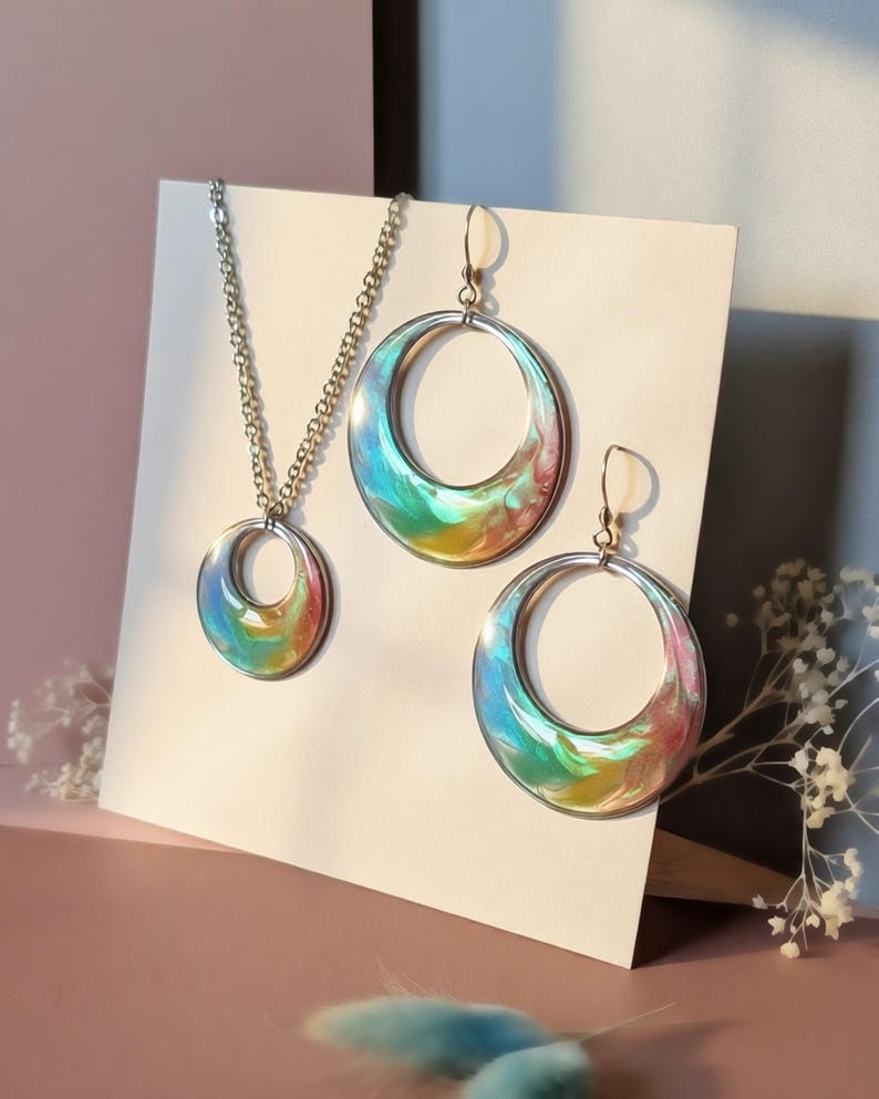 K&ouml;nnte beinhalten: Ein Schmuckset bestehend aus einer Halskette und zwei passenden Creolen. Jedes Schmuckst&uuml;ck verf&uuml;gt &uuml;ber einen kreisf&ouml;rmigen Metallrahmen mit einer wirbelnden, mehrfarbigen Harzeinlage in leuchtenden Blau-, Gr&uuml;n-, Gelb- und Rosat&ouml;nen.