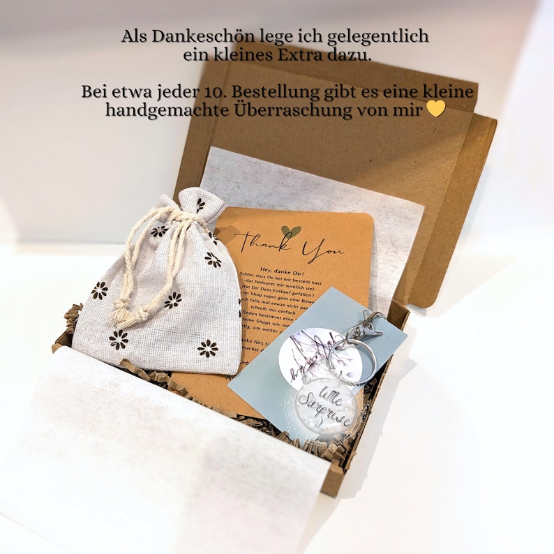 K&ouml;nnte beinhalten: Ein brauner Versandkarton aus Pappe mit einem kleinen beigen Beutel mit Kordelzug und schwarzem Blumenmuster, einer Dankeskarte sowie einem runden, durchsichtigen Schl&uuml;sselanh&auml;nger mit der Aufschrift Little Surprise an einem Metallring.