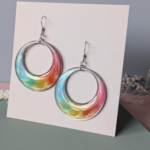 K&ouml;nnte beinhalten: Ein Paar kreisf&ouml;rmige Ohrringe mit einer wirbelnden, irisierenden Harzeinlage in leuchtenden Farbverl&auml;ufen aus Pink, Orange, Gelb und Blau. Die Ohrringe h&auml;ngen an silberfarbenen Metallhaken und sind auf einer wei&szlig;en Karte befestigt.