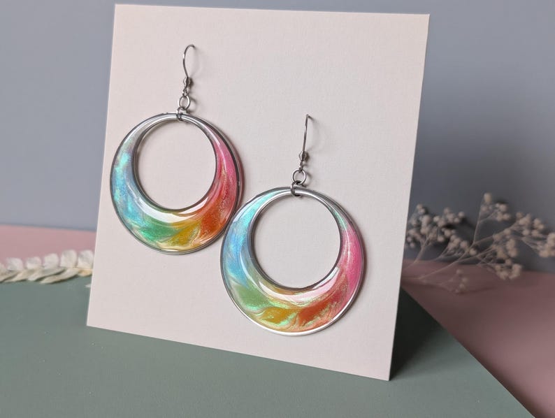 K&ouml;nnte beinhalten: Ein Paar kreisf&ouml;rmige Ohrringe mit einer wirbelnden, irisierenden Harzeinlage in leuchtenden Farbverl&auml;ufen aus Pink, Orange, Gelb und Blau. Die Ohrringe h&auml;ngen an silberfarbenen Metallhaken und sind auf einer wei&szlig;en Karte befestigt.