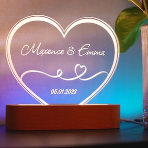 Peut inclure: Une plaque en acrylique en forme de cœur blanche avec les noms Maxence & Emma et la date 05.01.2023. La plaque est éclairée par une lumière blanche chaude et repose sur un socle en bois.