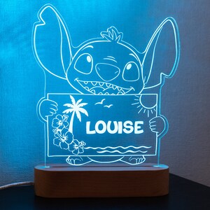 Peut inclure: Veilleuse LED bleue représentant un personnage de dessin animé tenant une pancarte avec le mot "LOUISE". La pancarte affiche une scène de plage tropicale avec un palmier, des fleurs et un soleil. La lumière est posée sur une base en bois.