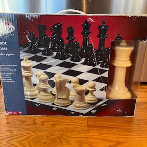 May include: A chess set with black and off-white pieces arranged on a checkered board. The set is in its original box, with some pieces still in their plastic packaging. The box has text in multiple languages.