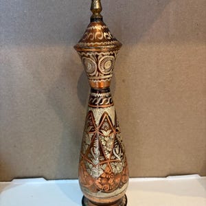 May include: A copper-colored vase with intricate floral and geometric patterns. The vase has a conical shape with a decorative lid and a base. The metal has a polished finish, and the design is etched into the surface.