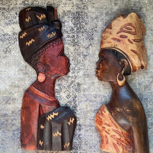 Könnte beinhalten: Zwei Holz-Wandskulpturen, die Profile afrikanischer Frauen darstellen. Die Skulpturen sind aus Holz geschnitzt und weisen komplizierte Details auf, darunter Kopfbedeckungen und Kleidung. Eine ist dunkelbraun mit goldenen Akzenten, die andere ist heller mit einem gemusterten Design.