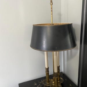 May include: A gold-toned table lamp with a black fabric lampshade. The lamp has three candle-like lights and a decorative finial on top.