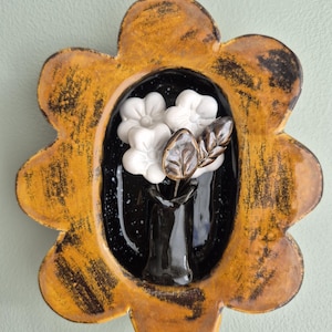 May include: A ceramic wall plaque shaped like a flower with a mustard yellow and black distressed finish. The centre features a glossy black oval inset containing a small black vase with three white flowers and two dark brown leaves.