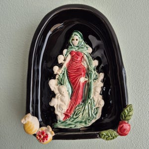 May include: A glossy black ceramic wall plaque featuring a central relief of a figure in a red dress draped in a green cloak, surrounded by small white skull-like shapes. The base is decorated with a small yellow fruit, a pomegranate, and a red rose.