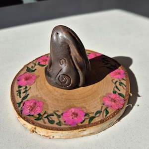 May include: A dark brown, abstract sculpture with a spiral design sits on a round wooden base. The base is decorated with pink flowers and green leaves. The sculpture is the focal point of the image.
