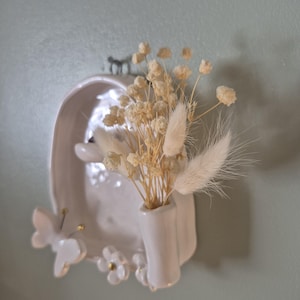 May include: A white ceramic wall-mounted vase featuring a sculpted butterfly and flower detail at the base. The vessel holds a small arrangement of dried cream-coloured baby's breath and fluffy bunny tail grass stems, creating a textured display.