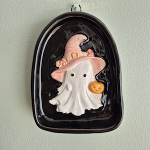 May include: A glossy black ceramic wall plaque featuring a raised, white ghost character wearing a light pink witch hat adorned with small flowers. The ghost holds a small orange pumpkin, and the entire piece has a rounded, arched shape.