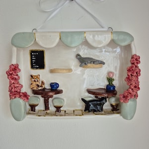 May include: A ceramic cat cafe ornament with a white and pale green awning. The interior features tables, cats, and flowers. A white ribbon is attached for hanging.