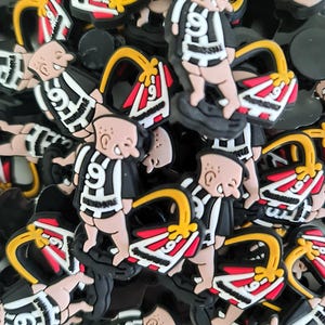 May include: A selection of shoe charms depicting a cartoon character in a black and white striped jersey with the number 9. The charms also include a shoe accessory with red, white, and yellow details. The charms are spread across a dark background.