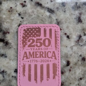 May include: A rectangular pink patch with rounded corners and stitched edges. The design features a distressed American flag graphic with the text 250 YEARS OF AMERICA and the dates 1776-2026 centered in a dark brown color on the pink surface.