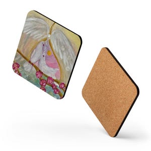 May include: Square coaster featuring a painting of two white doves on a branch with pink flowers. The background shows a yellow and pink sky with white angel wings. A second coaster with a cork surface is also visible.
