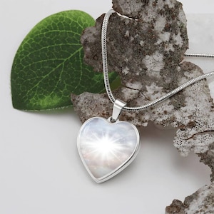 以下が含まれることがあります： 銀色のハート型のペンダントネックレス。中には白と銀の銀河のような画像が入っています。ネックレスは銀色のチェーンに付いており、木の板の上に置かれています。