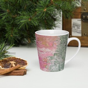 May include: A white ceramic latte mug with a pink and green abstract design. The mug has a tall, tapered shape and a handle.