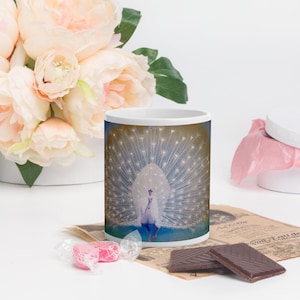 Pode incluir: Caneca de cerâmica branca com um pavão branco com as penas da cauda abertas. O pavão está sobre um fundo azul com um degradado amarelo e marrom.