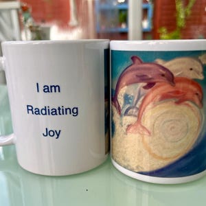 May include: White ceramic mug with a blue inscription that reads "I am radiating Joy". The other side of the mug features a colorful painting of three dolphins jumping over a wave.