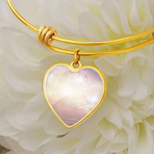 May include: A gold bangle bracelet with a heart-shaped charm. The charm features a pink and white image of clouds and a stairway to heaven.