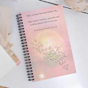 May include: A spiral-bound notebook with a pink cover featuring text that reads "New, Greater Horizons Beckon Me..." and "A Ta Eeya A Ku Ku Ku x 8." The cover also includes an abstract design in a darker shade.