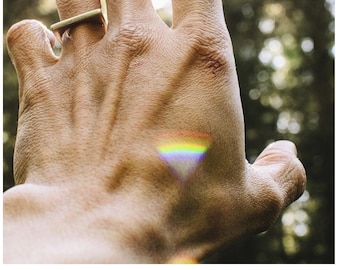 May include: A hand reaching towards the sun, with a gold ring on the index finger. A rainbow is visible in the light reflecting off the hand.