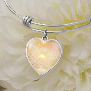 May include: A silver bangle bracelet with a heart-shaped charm. The charm has a gold mandala design with a white and pink background.