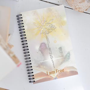 May include: Spiral-bound notebook with a celestial design. The cover features a golden starburst, angel wings, and an open book with the words "I am Free." The design has a watercolor effect with soft, blended colors. The notebook has a black spiral binding.